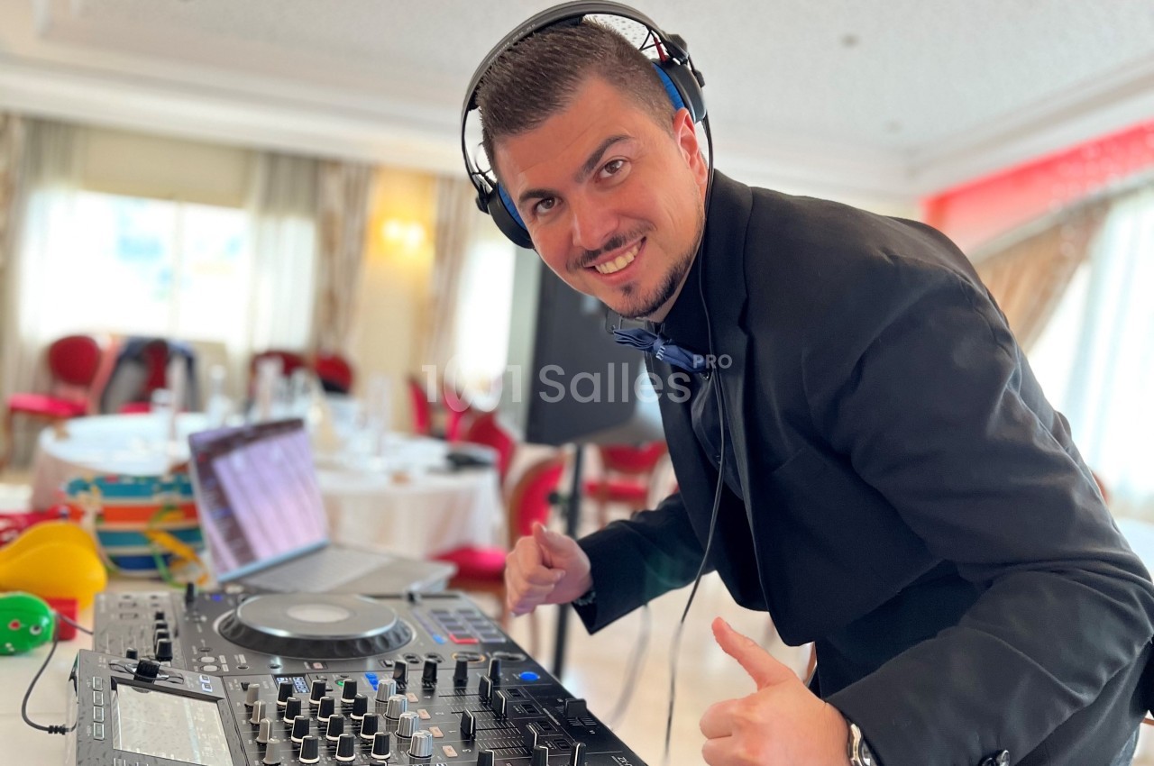 Un DJ souriant en costume mixe de la musique sur une table de mixage dans une salle lumineuse.