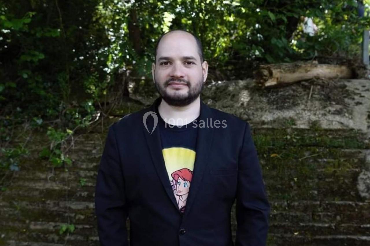 Homme debout devant un mur en pierre, portant une veste noire et un t-shirt imprimé, entouré de végétation.