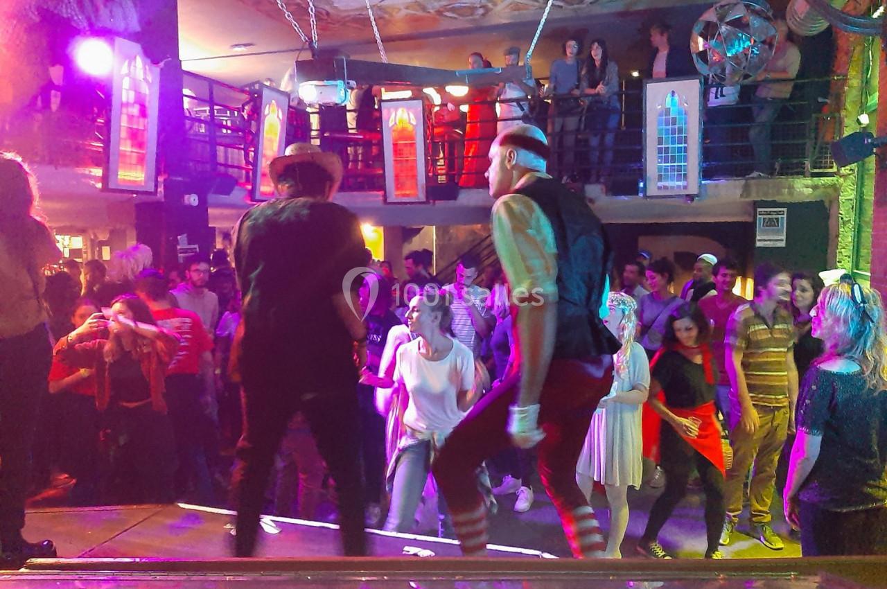 Des personnes dansent sur une scène et dans une salle animée, éclairée par des lumières colorées et décorée de vitraux.