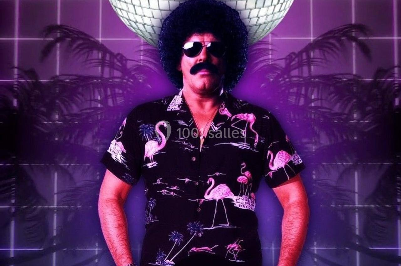 Homme en chemise à motifs et perruque afro tenant un disque vinyle devant une boule disco lumineuse.