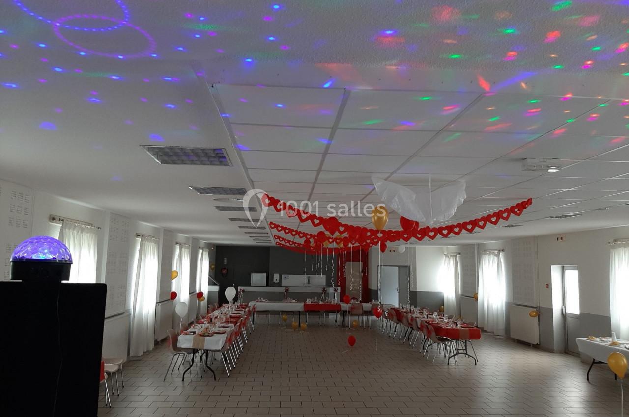 Salle décorée pour une fête avec guirlandes rouges, ballons et tables dressées, éclairée par des lumières colorées.