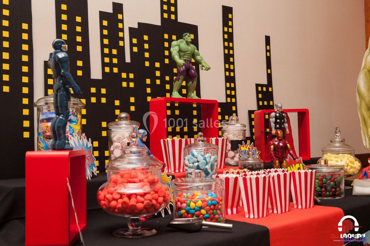 Table décorée sur le thème des super-héros avec bonbons, popcorn et figurines devant un décor de ville stylisé.