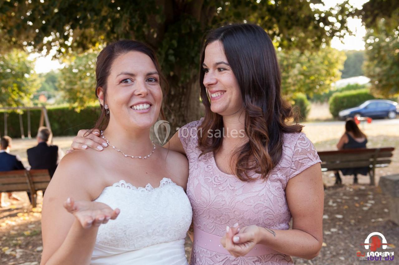 Deux femmes souriantes en tenue élégante, dont une en robe de mariée, posent devant un arbre dans un parc.
