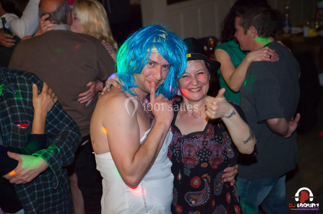 Un homme déguisé en robe blanche et perruque bleue pose avec une femme souriante lors d'une fête animée.