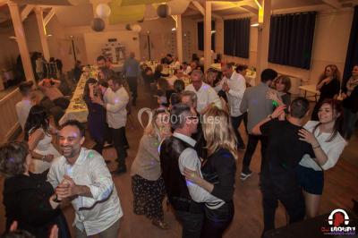 Des personnes dansent en ligne dans une salle décorée de ballons, avec des tables dressées et des convives en arrière-plan.