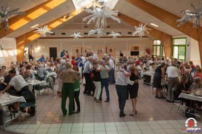 Des personnes dansent en ligne dans une salle décorée de ballons, avec des tables dressées et des convives en arrière-plan.