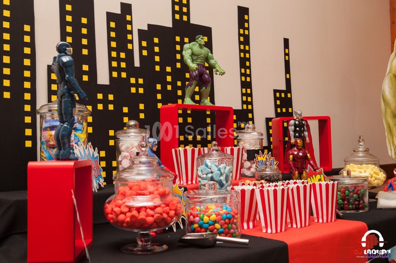 Table décorée sur le thème des super-héros avec bonbons, popcorn et figurines devant un décor de ville stylisé.