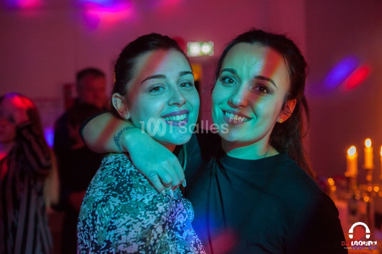 Deux femmes souriantes posent ensemble dans une ambiance festive avec des lumières colorées en arrière-plan.
