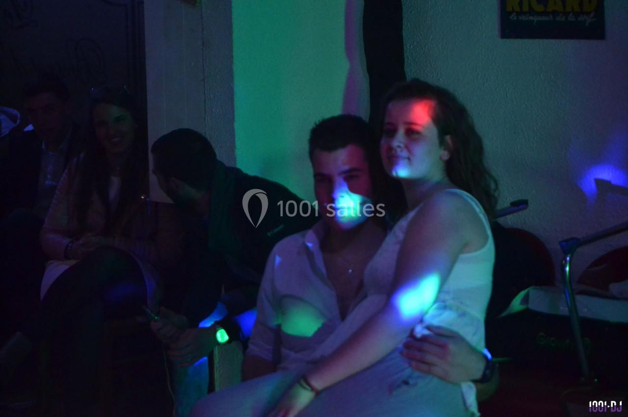 Un couple assis dans une ambiance tamisée avec des lumières colorées, entouré de personnes en arrière-plan.
