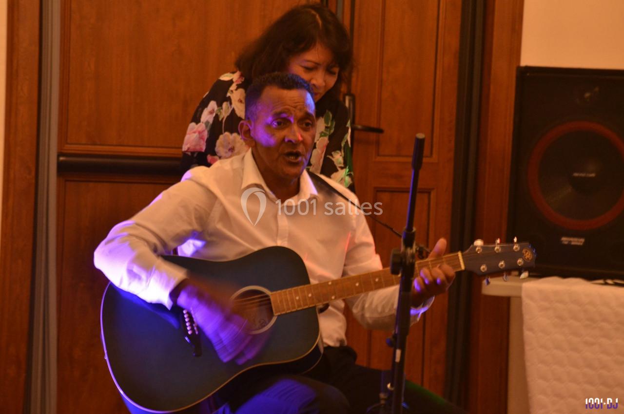 Un homme joue de la guitare acoustique en chantant, accompagné d'une femme debout derrière lui.