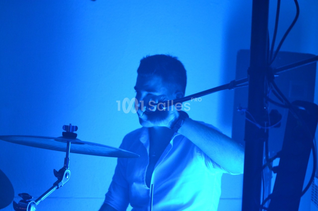 Un batteur en chemise blanche joue sur une batterie sous un éclairage bleu intense.