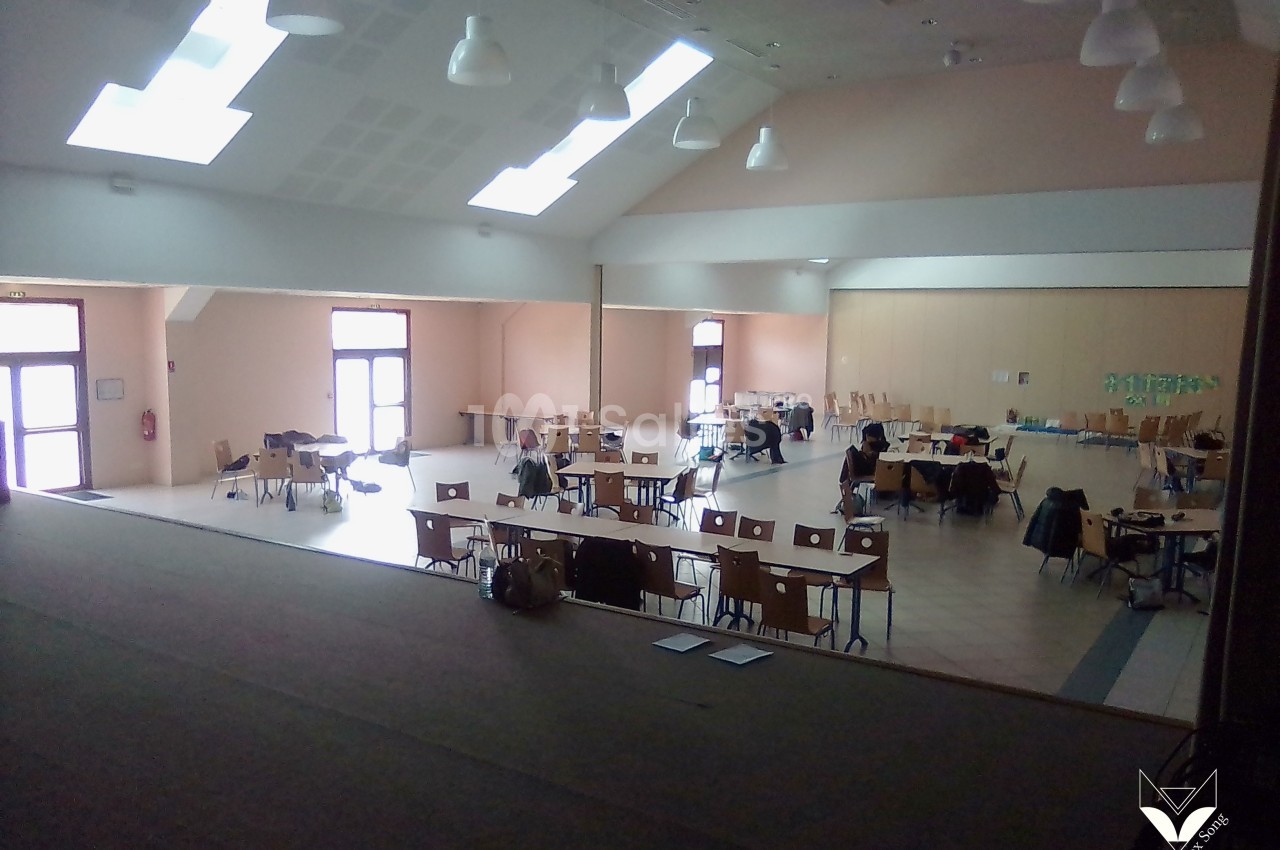Salle lumineuse avec des tables et des chaises dispersées, quelques personnes assises dans un espace intérieur spacieux.