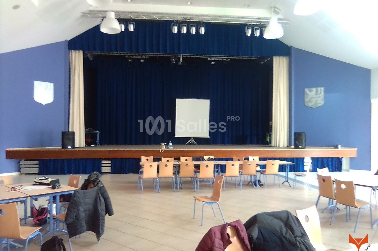 Salle polyvalente avec scène surélevée, rideaux bleus, écran blanc, tables et chaises disposées en avant-plan.