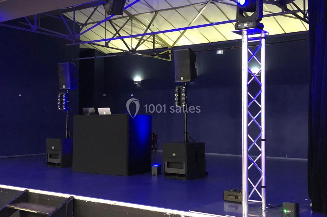 Scène avec une table de mixage, haut-parleurs, éclairage bleu et structure métallique dans une salle sombre.