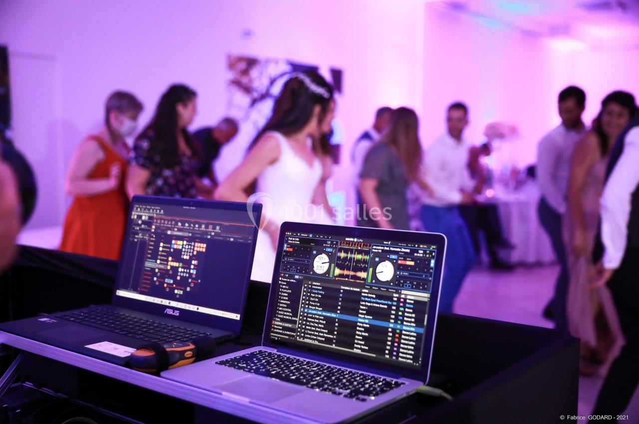 Deux ordinateurs affichant des logiciels de mixage musical, en arrière-plan des invités dansent dans une salle éclairée.