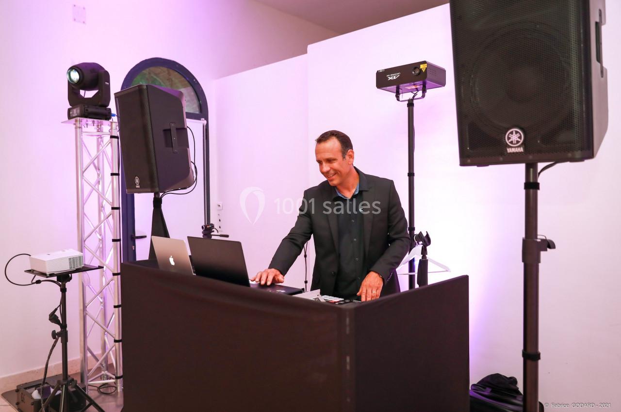 Un DJ souriant mixe de la musique derrière une table équipée d'ordinateurs et d'enceintes dans une salle éclairée.