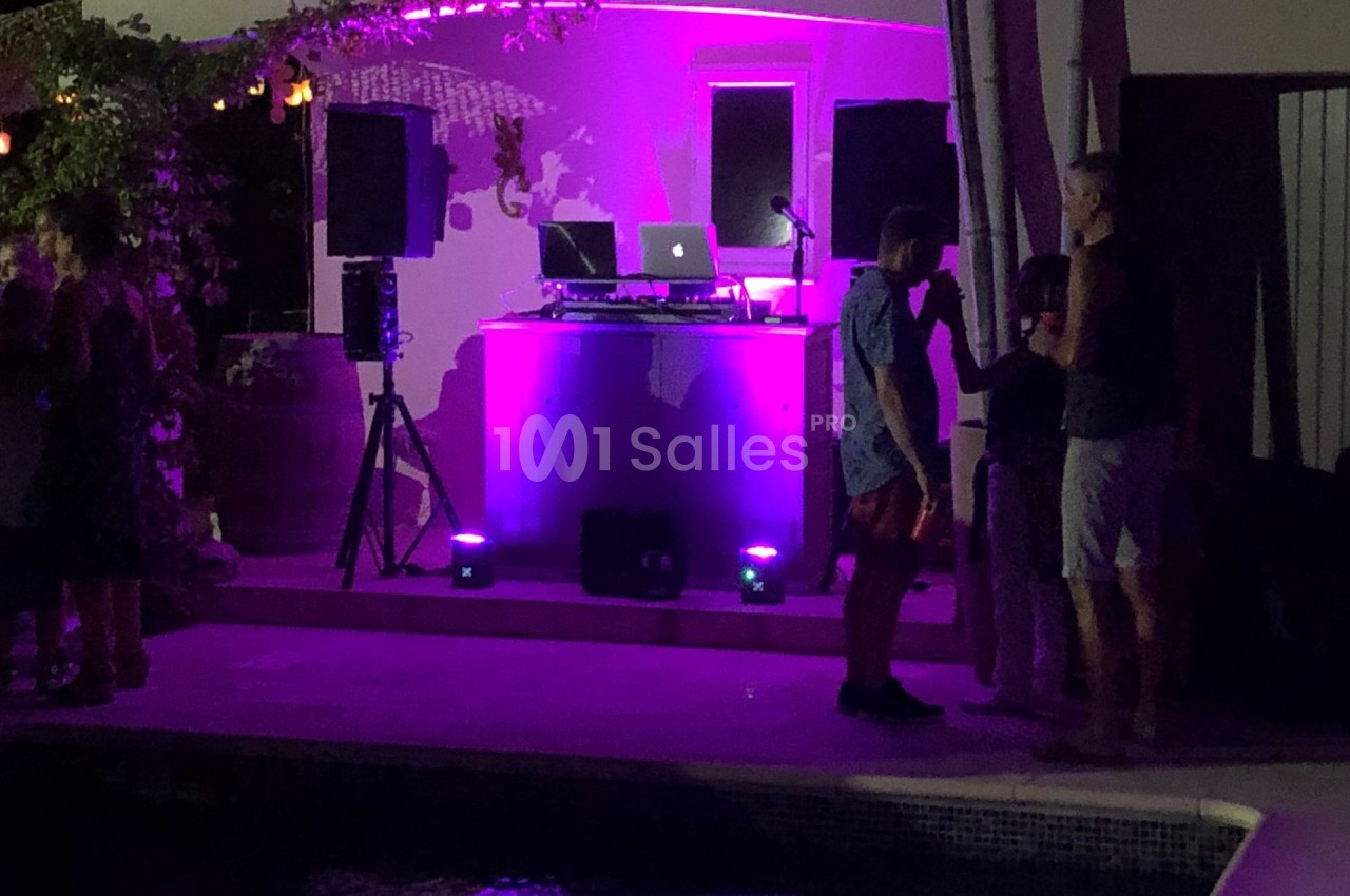 Scène de soirée en extérieur avec éclairage violet, une table de mixage et des personnes discutant près d'une piscine.