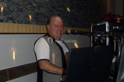 Miniature DJ Buchy (Seine-Maritime) - Djmanfred - Animmix #16 Homme souriant debout devant un micro, portant une chemise noire avec un gilet rayé, fond marron.