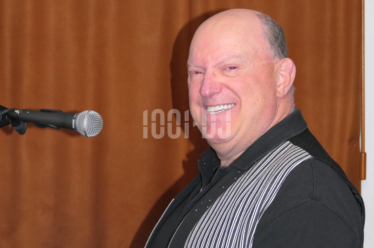Homme souriant debout devant un micro, portant une chemise noire avec un gilet rayé, fond marron.