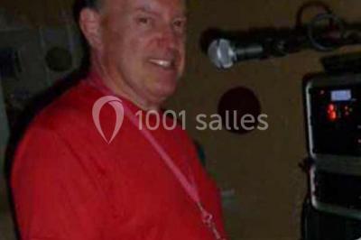 Miniature DJ Buchy (Seine-Maritime) - Djmanfred - Animmix #3 Homme souriant debout devant un micro, portant une chemise noire avec un gilet rayé, fond marron.