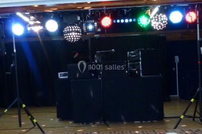 Miniature DJ Buchy (Seine-Maritime) - Djmanfred - Animmix #6 Homme souriant debout devant un micro, portant une chemise noire avec un gilet rayé, fond marron.