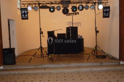 Miniature DJ Buchy (Seine-Maritime) - Djmanfred - Animmix #13 Homme souriant debout devant un micro, portant une chemise noire avec un gilet rayé, fond marron.