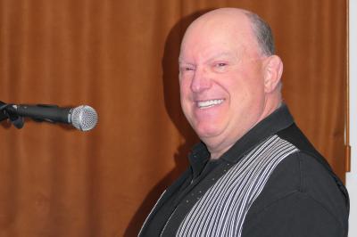 Miniature DJ Buchy (Seine-Maritime) - Djmanfred - Animmix #19 Homme souriant debout devant un micro, portant une chemise noire avec un gilet rayé, fond marron.