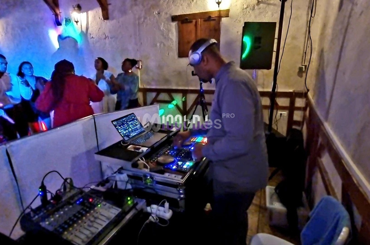 Un DJ mixe de la musique sur une console dans une salle avec des lumières colorées et des personnes dansant en arrière-plan.