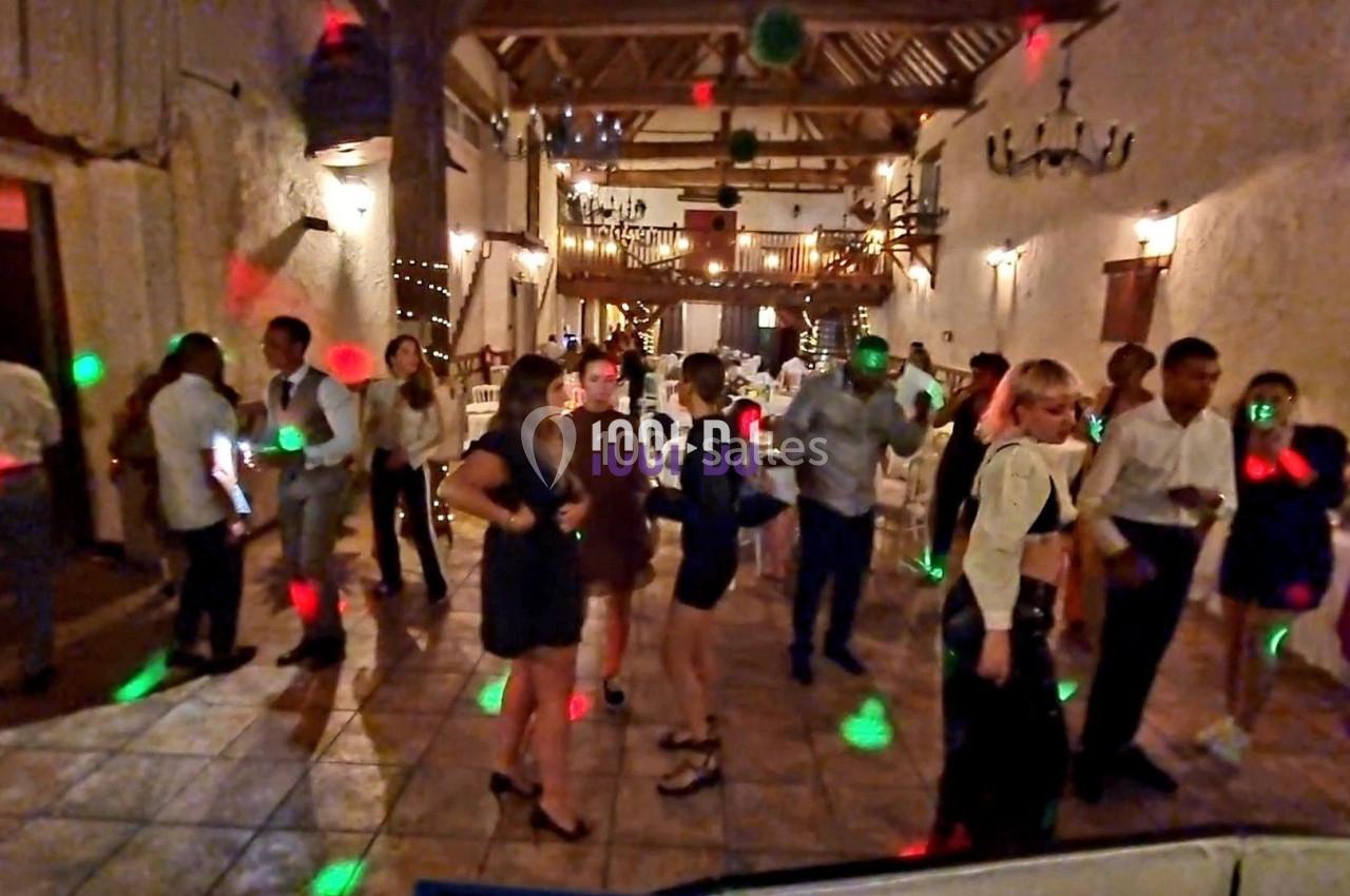 Des personnes dansent dans une salle de réception éclairée par des lumières colorées, avec un décor rustique en arrière-plan.