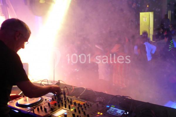 Un DJ mixe sur une console dans une ambiance lumineuse et enfumée, avec des personnes dansant en arrière-plan.