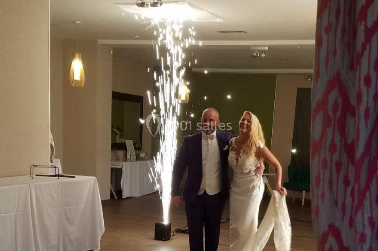 Un couple en tenue de mariage entre dans une salle éclairée, accompagné d'effets pyrotechniques en arrière-plan.
