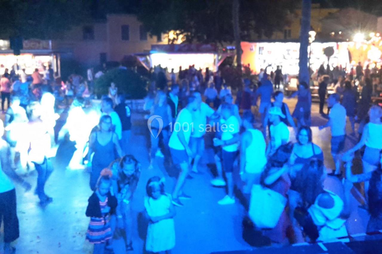 Des personnes dansent et se rassemblent en soirée sur une place éclairée par des lumières colorées, avec des stands en…