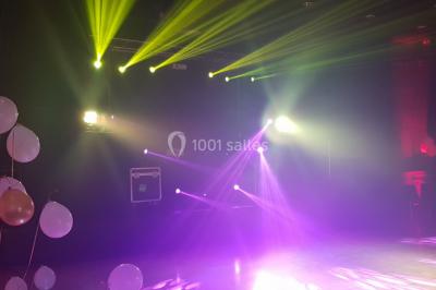 Salle avec éclairage de scène coloré, piste de danse illuminée, tables sur les côtés et matériel audio en arrière-plan.