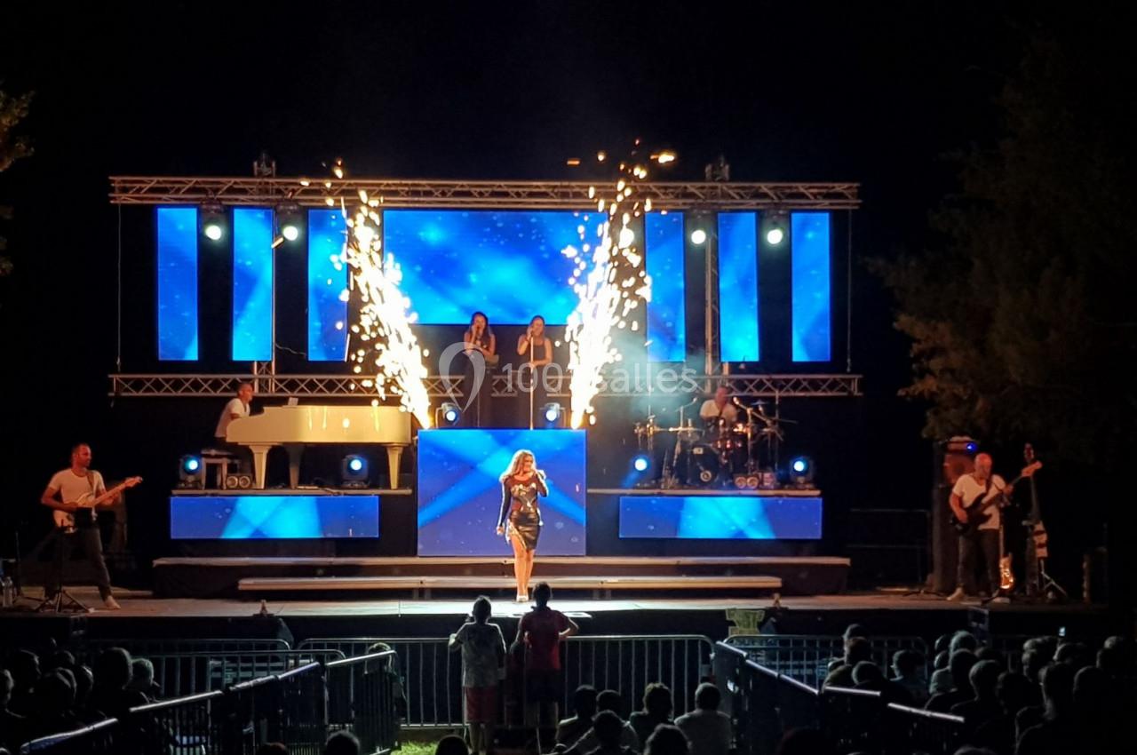 Scène de concert nocturne avec une chanteuse au centre, des musiciens, des écrans lumineux et des effets pyrotechniques.