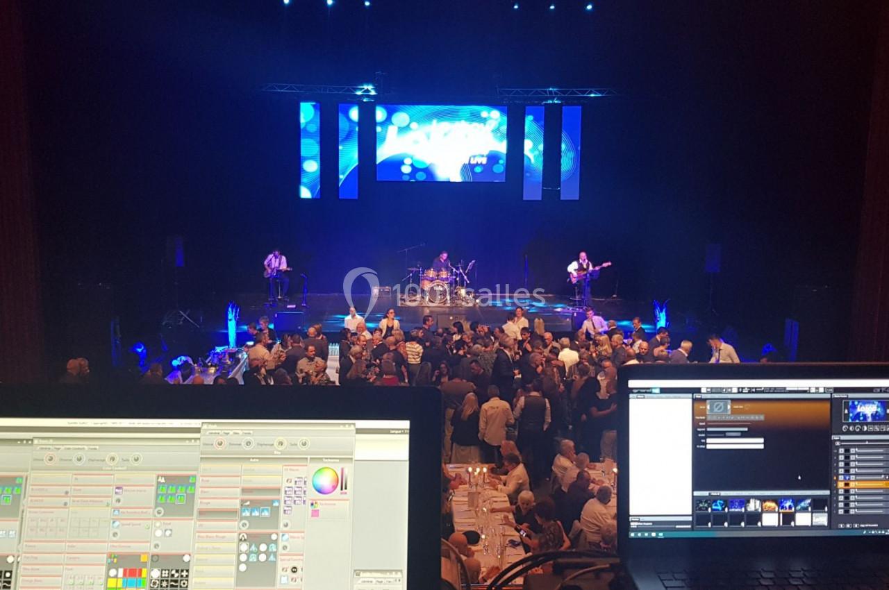 Vue depuis une régie technique sur un concert avec musiciens sur scène, public debout et écrans lumineux en arrière-plan.