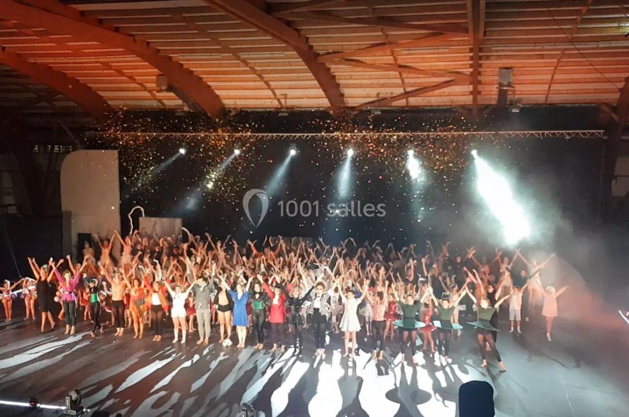 Un grand groupe de personnes sur scène sous des projecteurs et des confettis, dans une salle avec une structure en bois.