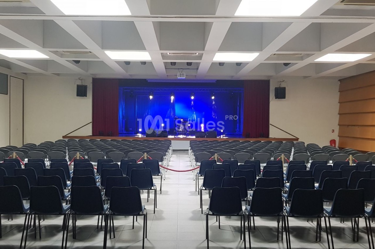 Salle de spectacle vide avec des rangées de chaises noires face à une scène éclairée en bleu.