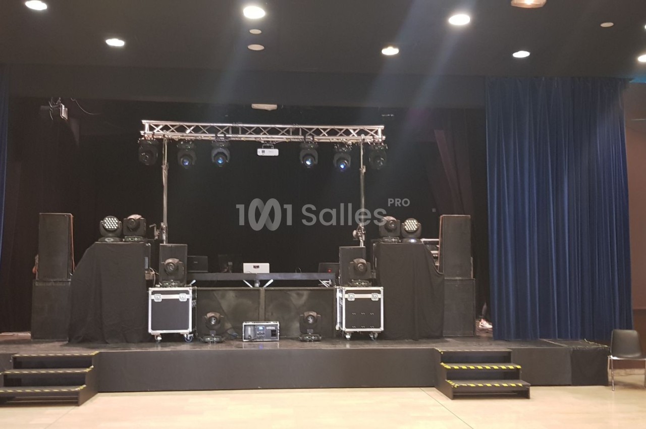 Scène équipée de matériel de sonorisation et d'éclairage, entourée de rideaux bleus dans une salle de spectacle.