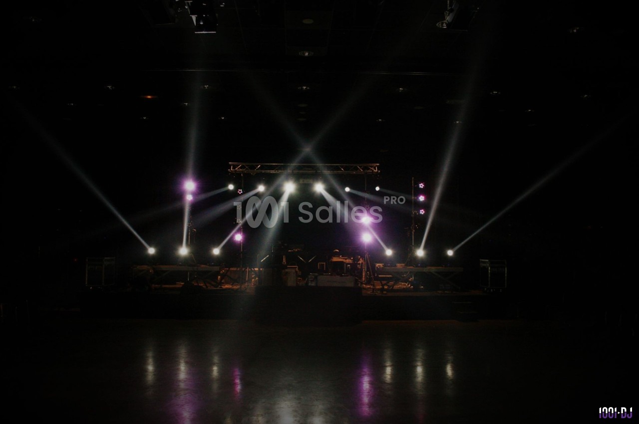 Scène de concert vide éclairée par des projecteurs, avec des équipements et instruments visibles dans une salle sombre.