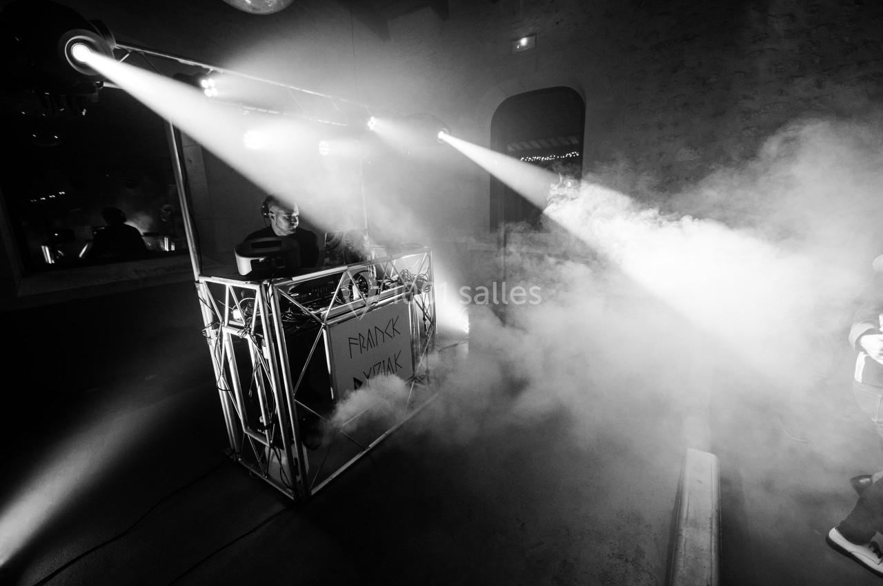 Un DJ joue de la musique sur une scène éclairée par des projecteurs, entouré de fumée dans une ambiance nocturne.