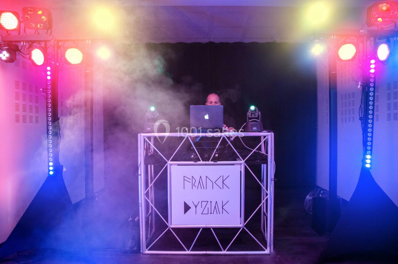 Un DJ mixe derrière une table éclairée par des lumières colorées et entourée de fumée.