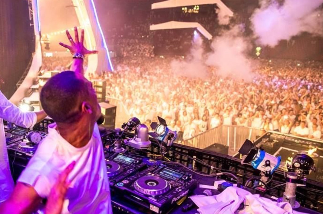 Un DJ anime une foule dense lors d'un concert en plein air, avec des lumières et de la fumée sur scène.