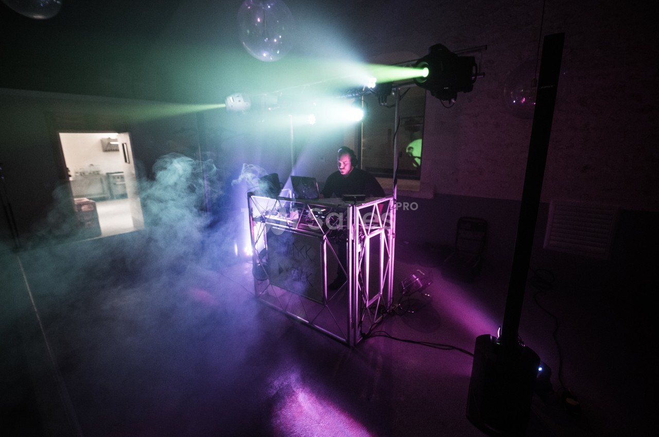 Un DJ mixe sur une scène éclairée par des projecteurs colorés, entouré de fumée dans une ambiance nocturne.