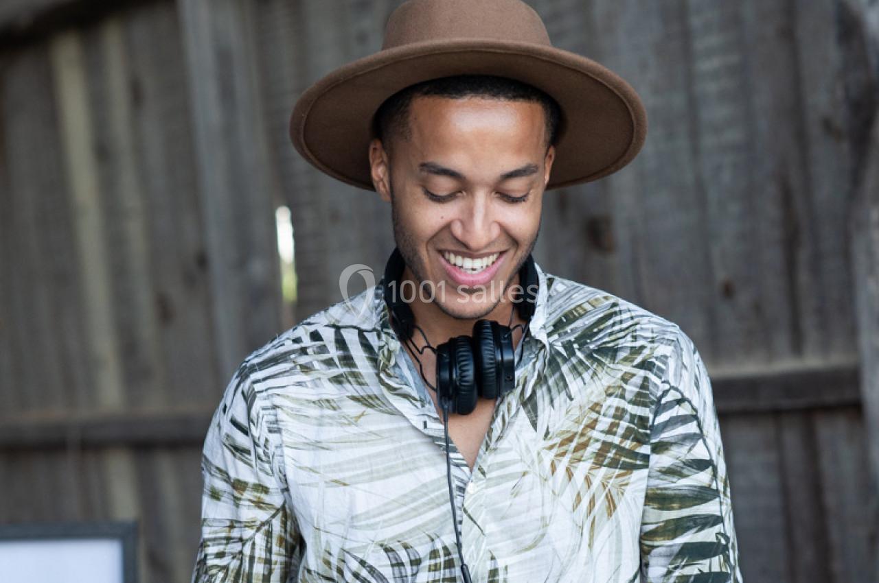 Homme souriant portant un chapeau marron, une chemise à motif tropical et un casque audio autour du cou.