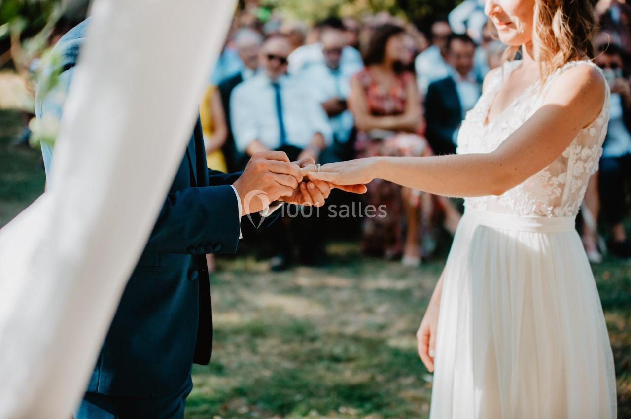 Un homme en costume passe une alliance au doigt d'une femme en robe blanche lors d'une cérémonie en extérieur.