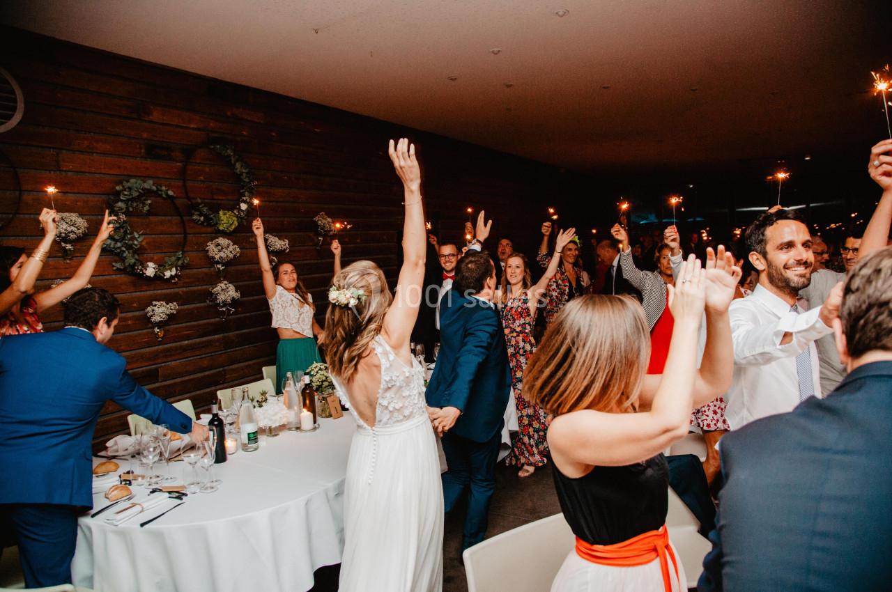 Des invités dansent et lèvent les bras lors d'une fête dans une salle décorée avec des lumières et des fleurs.