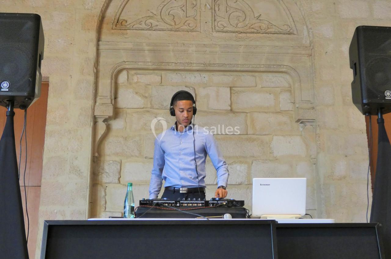 Un DJ mixe de la musique sur une table équipée de matériel audio, dans une salle en pierre avec des enceintes.