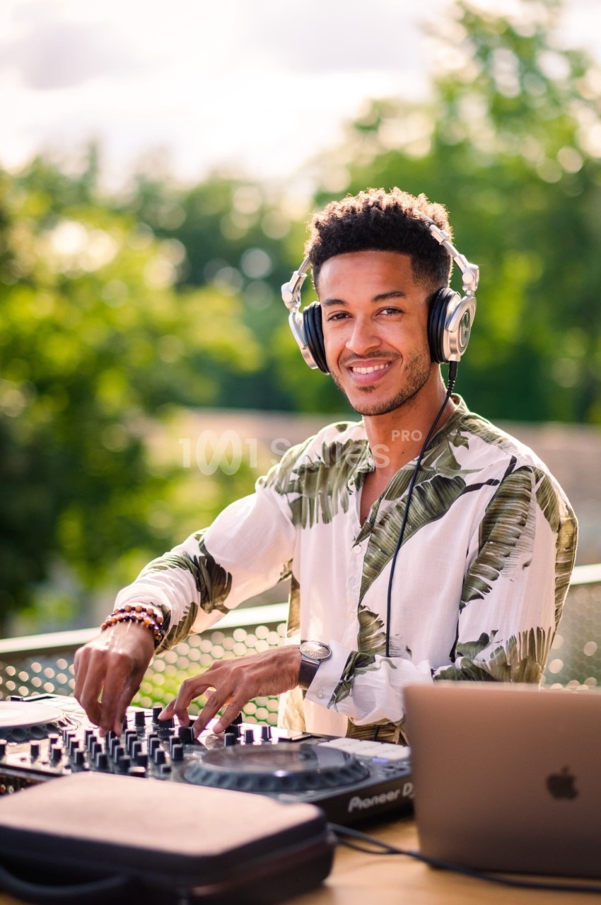 Un DJ souriant mixe de la musique en extérieur, entouré de verdure, avec un ordinateur portable à ses côtés.