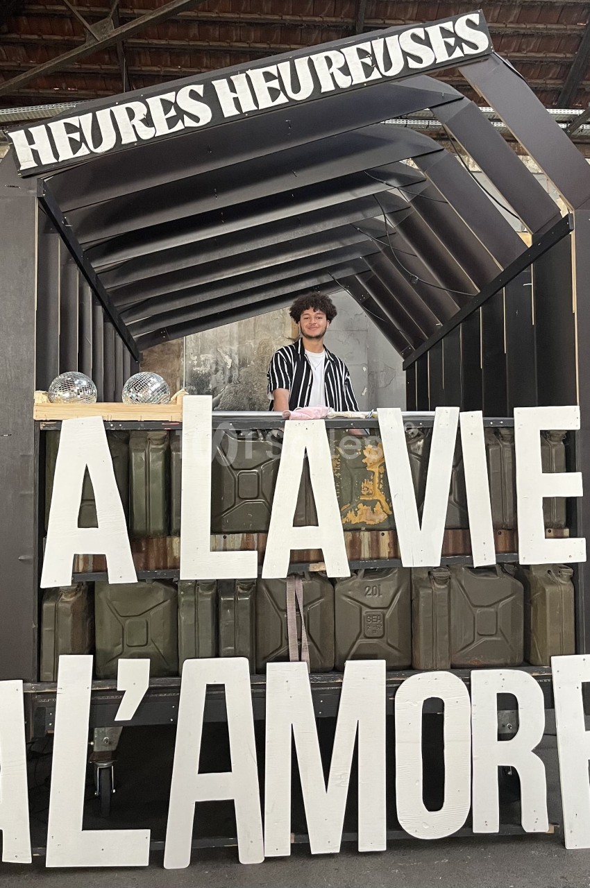 Stand décoratif avec une personne souriante derrière un comptoir, affichant les mots ’À la vie, à l'amore’.