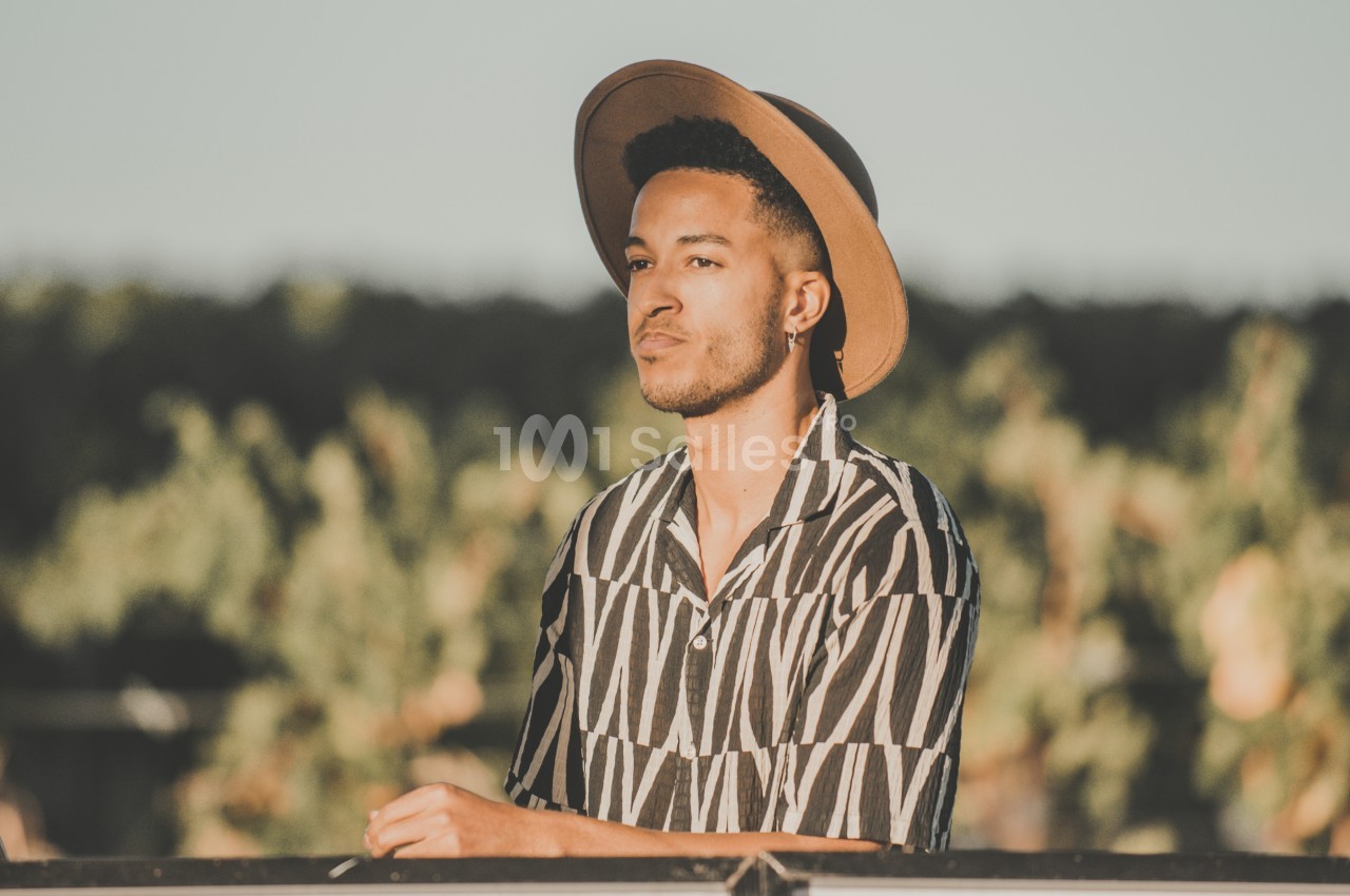 Un homme portant un chapeau et une chemise à motifs se tient devant un ordinateur en extérieur, avec des arbres en arrière…